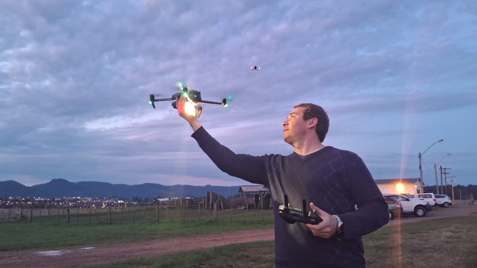 A foto mostra um homem em um campo aberto, no fim da tarde, segurando um controle remoto de drone com a mão esquerda enquanto lança o equipamento com a mão direita. O drone, iluminado por luzes verdes, está em voo próximo à sua mão. O céu está parcialmente nublado, com tons azulados e rosados, enquanto ao fundo aparecem cercas, casas rurais, veículos estacionados e a silhueta de montanhas.