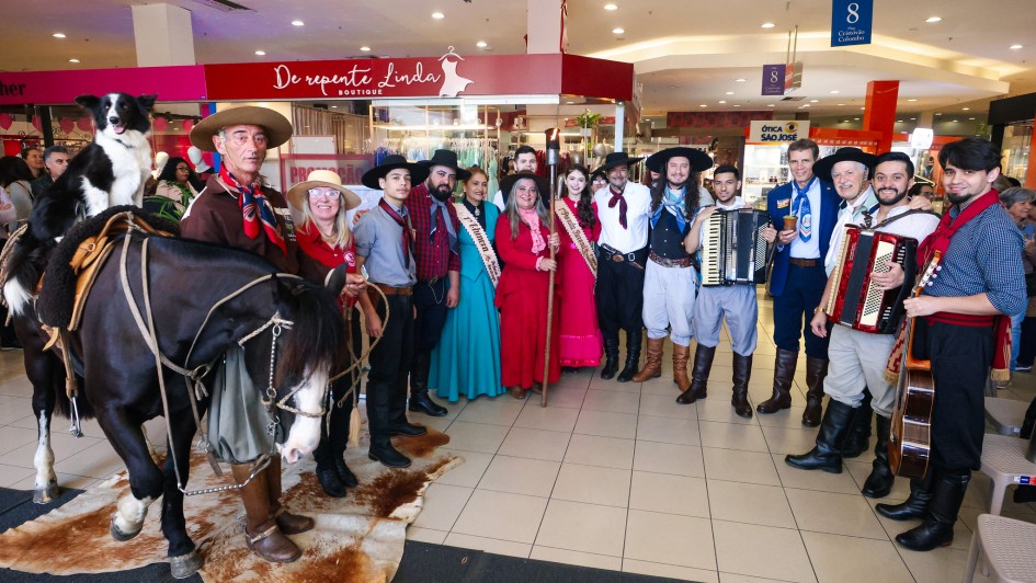 A imagem mostra um grupo de pessoas reunidas em um shopping center, vestidas com trajes típicos da cultura gaúcha, tradicional do sul do Brasil. No canto esquerdo, há um homem montado a cavalo com um cachorro preto e branco sentado na parte traseira da sela. O cavalo está parado sobre um tapete feito de couro bovino. Ao redor, estão homens e mulheres com bombachas, lenços, vestidos longos e chapéus, formando uma fila para a foto.

Entre o grupo, há três músicos segurando instrumentos tradicionais da música gaúcha: dois acordeons e um violão. Duas mulheres usam faixas, indicando que são representantes de algum tipo de concurso cultural ou tradição (como prendas ou embaixatrizes). Ao fundo, vemos vitrines de lojas e algumas placas indicativas do shopping, como “De Repente Linda Boutique” e “Ótica São José”.

O ambiente é festivo, e o grupo parece estar participando de uma celebração cultural ou evento tradicionalista dentro do shopping.