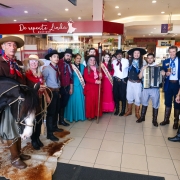 A imagem mostra um grupo de pessoas reunidas em um shopping center, vestidas com trajes típicos da cultura gaúcha, tradicional do sul do Brasil. No canto esquerdo, há um homem montado a cavalo com um cachorro preto e branco sentado na parte traseira da sela. O cavalo está parado sobre um tapete feito de couro bovino. Ao redor, estão homens e mulheres com bombachas, lenços, vestidos longos e chapéus, formando uma fila para a foto.

Entre o grupo, há três músicos segurando instrumentos tradicionais da música gaúcha: dois acordeons e um violão. Duas mulheres usam faixas, indicando que são representantes de algum tipo de concurso cultural ou tradição (como prendas ou embaixatrizes). Ao fundo, vemos vitrines de lojas e algumas placas indicativas do shopping, como “De Repente Linda Boutique” e “Ótica São José”.

O ambiente é festivo, e o grupo parece estar participando de uma celebração cultural ou evento tradicionalista dentro do shopping.