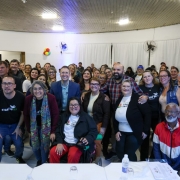 Descrição da imagem: Um grupo numeroso de pessoas está reunido em um salão iluminado, posando para uma foto coletiva. Na frente, algumas pessoas estão sentadas e outras de pé, incluindo mulheres e homens de diferentes idades e estilos. Algumas usam camisetas com os logos “Clube RS” e “Famílias Gaúchas”, sugerindo participação em um evento comunitário ou institucional. Há também uma mulher em cadeira de rodas no centro da primeira fila, sorridente. Ao fundo, outras pessoas formam uma segunda e terceira fileira, todas olhando para a câmera. O ambiente tem paredes brancas, cortinas e ventiladores fixados, além de algumas bexigas coloridas na decoração lateral. O clima é de confraternização e inclusão.