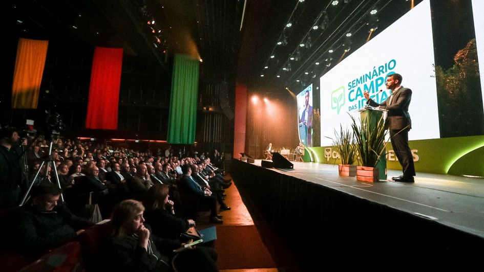 Auditório lotado durante um evento oficial. No palco, um homem de terno fala em um púlpito iluminado, com projeção de sua imagem em uma grande tela ao fundo. Na tela aparece também o logotipo do evento com o texto “Seminário Campo das Ideias”. O cenário do palco é decorado com plantas em vasos. Na plateia, muitas pessoas assistem atentamente à apresentação. Ao fundo do auditório, pendem do teto três grandes faixas verticais nas cores amarela, vermelha e verde, lembrando a bandeira do Rio Grande do Sul.