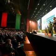 Auditório lotado durante um evento oficial. No palco, um homem de terno fala em um púlpito iluminado, com projeção de sua imagem em uma grande tela ao fundo. Na tela aparece também o logotipo do evento com o texto “Seminário Campo das Ideias”. O cenário do palco é decorado com plantas em vasos. Na plateia, muitas pessoas assistem atentamente à apresentação. Ao fundo do auditório, pendem do teto três grandes faixas verticais nas cores amarela, vermelha e verde, lembrando a bandeira do Rio Grande do Sul.