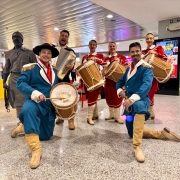 A imagem mostra um grupo de artistas trajando roupas típicas gaúchas em apresentação cultural dentro do Aeroporto Salgado Filho. Dois homens vestidos de bombacha azul, lenço vermelho e botas seguram tambores e estão ajoelhados em destaque. Atrás deles, quatro mulheres de vestido vermelho tocam instrumentos, e um homem toca acordeom. Ao fundo, um artista caracterizado como estátua humana também participa da cena. O ambiente moderno do saguão contrasta com a tradição, criando clima festivo de celebração do Mês Farroupilha.
