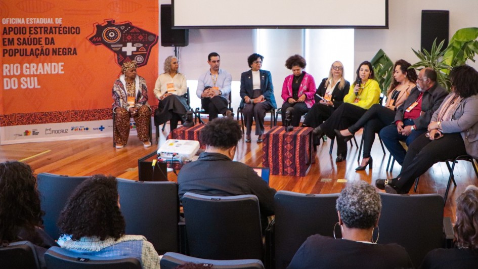 A imagem mostra um painel de discussão com 12 pessoas sentadas em círculo, participando da Oficina Estadual de Apoio Estratégico em Saúde da População Negra, realizada no Rio Grande do Sul. O fundo apresenta um banner laranja com o nome do evento, uma ilustração estilizada do mapa do Brasil e símbolos culturais africanos.

Os participantes no palco são diversos, incluindo mulheres e homens, a maioria pessoas negras, vestindo roupas formais ou culturais. Uma das participantes segura um microfone e fala, enquanto os demais escutam atentamente. Na frente do palco, há algumas cadeiras ocupadas pelo público, um projetor e um notebook sendo operado por uma pessoa de costas.

O ambiente é interno, com piso de madeira, plantas decorativas e iluminação natural vinda das janelas. O clima é de escuta, diálogo e construção coletiva sobre políticas de saúde voltadas à população negra.