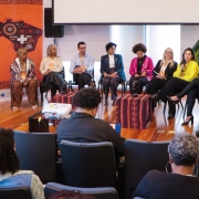 A imagem mostra um painel de discussão com 12 pessoas sentadas em círculo, participando da Oficina Estadual de Apoio Estratégico em Saúde da População Negra, realizada no Rio Grande do Sul. O fundo apresenta um banner laranja com o nome do evento, uma ilustração estilizada do mapa do Brasil e símbolos culturais africanos.

Os participantes no palco são diversos, incluindo mulheres e homens, a maioria pessoas negras, vestindo roupas formais ou culturais. Uma das participantes segura um microfone e fala, enquanto os demais escutam atentamente. Na frente do palco, há algumas cadeiras ocupadas pelo público, um projetor e um notebook sendo operado por uma pessoa de costas.

O ambiente é interno, com piso de madeira, plantas decorativas e iluminação natural vinda das janelas. O clima é de escuta, diálogo e construção coletiva sobre políticas de saúde voltadas à população negra.