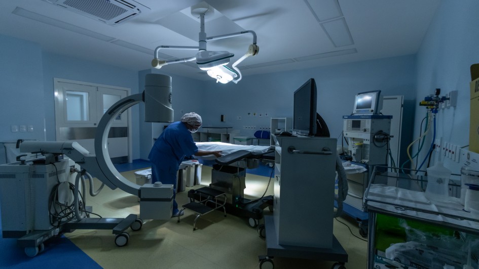 Foto de uma sala cirúrgica bem equipada, com luz de tom azul. Uma pessoa com trajes hospitalares azuis aparece ao centro, arrumando um leito cirúrgico.