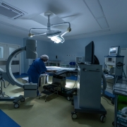 Foto de uma sala cirúrgica bem equipada, com luz de tom azul. Uma pessoa com trajes hospitalares azuis aparece ao centro, arrumando um leito cirúrgico.