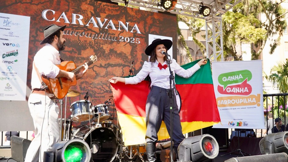 A foto mostra apresentação no palco da Caravana Farroupilha 2025, instalado ao ar livre no centro de Porto Alegre. Uma mulher com trajes gaúchos tradicionais, chapéu preto e lenço vermelho, segura a bandeira do Rio Grande do Sul enquanto canta ao microfone. Ao lado, um homem toca violão, e ao fundo há bateria e instrumentos. O painel atrás exibe “Caravana Farroupilha 2025”.