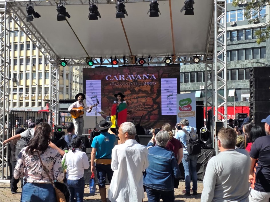 A foto mostra o público reunido diante do palco da Caravana Farroupilha 2025, montado no centro de Porto Alegre. Em cena, uma cantora segura a bandeira do Rio Grande do Sul enquanto se apresenta ao lado de um músico com violão. No painel de fundo está escrito “Caravana Farroupilha 2025”, ladeado por logomarcas de apoiadores. A plateia, de pé, assiste atentamente, compondo uma atmosfera de festa popular e valorização da cultura tradicionalista em meio ao cenário urbano.