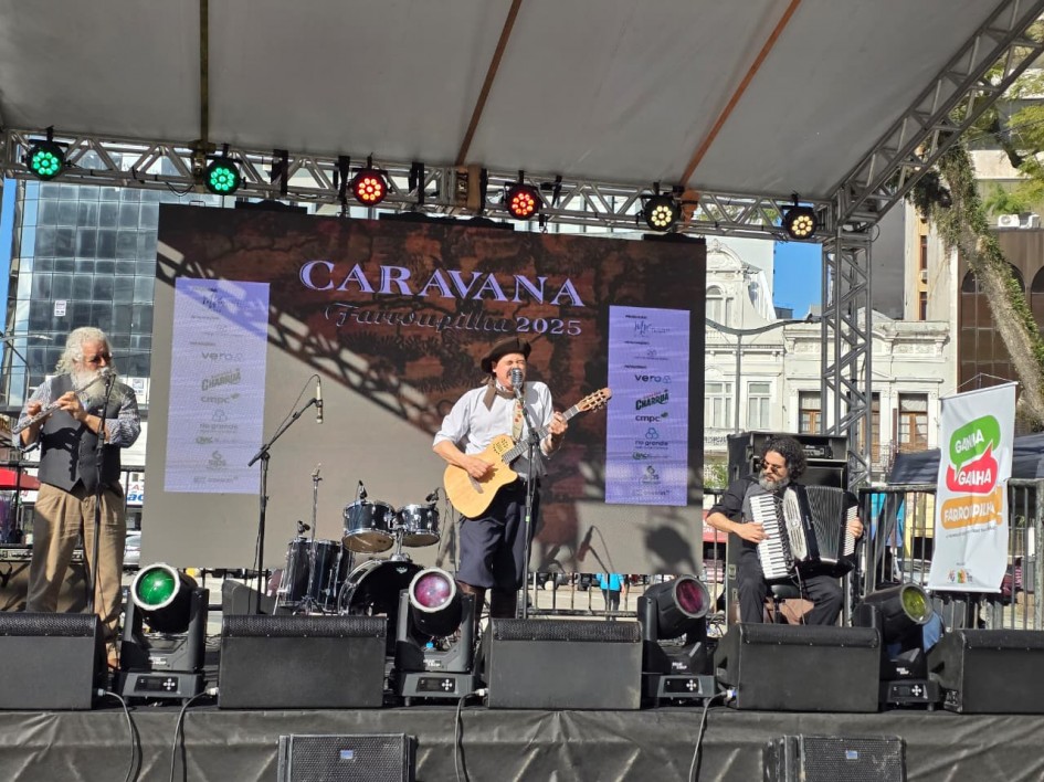 A imagem mostra três músicos se apresentando no palco da Caravana Farroupilha 2025. À esquerda, um homem de cabelos brancos toca flauta; ao centro, outro músico canta e toca violão; à direita, um acordeonista acompanha sentado. O painel de fundo exibe “Caravana Farroupilha 2025” sobre mapa estilizado. No canto, banner com a marca “Ganha Ganha Farroupilha”.