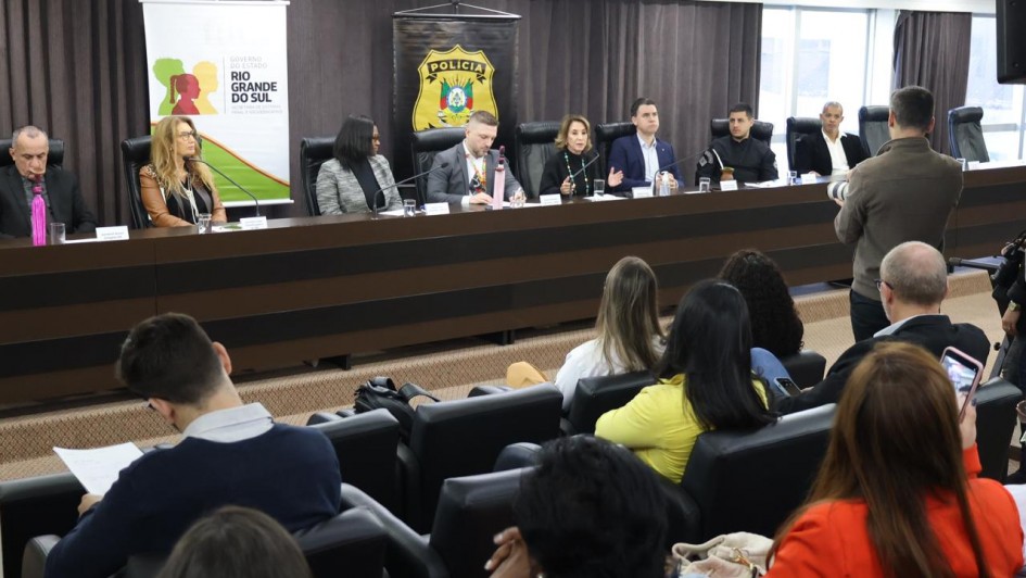A imagem mostra uma mesa de autoridades em um auditório, durante o primeiro encontro estadual de enfrentamento ao racismo no sistema prisional gaúcho. Diversos representantes estão sentados lado a lado, com microfones à frente e público acompanhando. Atrás da mesa, banners exibem os logotipos do Governo do Estado do Rio Grande do Sul e da Polícia Civil.