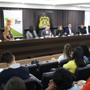 A imagem mostra uma mesa de autoridades em um auditório, durante o primeiro encontro estadual de enfrentamento ao racismo no sistema prisional gaúcho. Diversos representantes estão sentados lado a lado, com microfones à frente e público acompanhando. Atrás da mesa, banners exibem os logotipos do Governo do Estado do Rio Grande do Sul e da Polícia Civil.