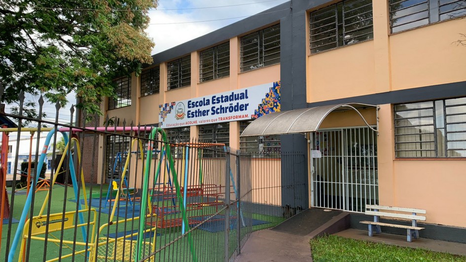 A imagem mostra a fachada da Escola Estadual Esther Schröder, um prédio de dois andares com paredes em tons de bege e cinza, localizado atrás de um pátio com brinquedos coloridos, como balanços e escorregadores, indicativos de um espaço voltado para crianças.

Acima da entrada principal da escola há uma grande faixa com o nome completo da instituição e o lema:
"Educação que acolhe, valores que transformam."

A entrada é protegida por um portão de grade branca com uma pequena cobertura metálica curva. À frente do prédio há uma cerca de ferro e uma calçada que leva até uma rampa de acessibilidade. Também há um pequeno banco pintado de branco e azul, posicionado ao lado da entrada. Uma árvore grande e frondosa está visível à esquerda, projetando sombra sobre o pátio.

O ambiente parece limpo, organizado e acolhedor, transmitindo a sensação de um espaço seguro e dedicado à educação infantil e fundamental.