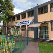 A imagem mostra a fachada da Escola Estadual Esther Schröder, um prédio de dois andares com paredes em tons de bege e cinza, localizado atrás de um pátio com brinquedos coloridos, como balanços e escorregadores, indicativos de um espaço voltado para crianças.

Acima da entrada principal da escola há uma grande faixa com o nome completo da instituição e o lema:
"Educação que acolhe, valores que transformam."

A entrada é protegida por um portão de grade branca com uma pequena cobertura metálica curva. À frente do prédio há uma cerca de ferro e uma calçada que leva até uma rampa de acessibilidade. Também há um pequeno banco pintado de branco e azul, posicionado ao lado da entrada. Uma árvore grande e frondosa está visível à esquerda, projetando sombra sobre o pátio.

O ambiente parece limpo, organizado e acolhedor, transmitindo a sensação de um espaço seguro e dedicado à educação infantil e fundamental.