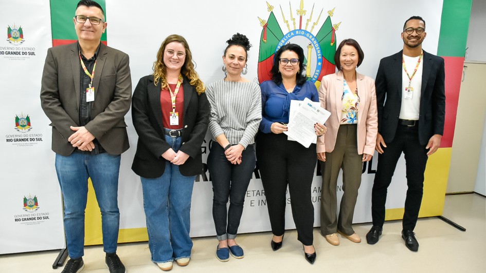 Seis pessoas estão alinhadas em pé, posando para a foto diante de um painel institucional do Governo do Estado do Rio Grande do Sul, que traz o brasão oficial no centro e as cores verde, vermelho e amarelo nas laterais. Da esquerda para a direita:
Um homem de óculos, cabelo curto escuro, veste blazer marrom, camisa preta e jeans.
Uma mulher de cabelos ondulados loiros, óculos e sorriso, veste blazer preto, blusa vinho e calça jeans larga.
Uma mulher de cabelos presos em coque alto, veste blusa cinza listrada e calça preta.
Uma mulher de cabelos escuros soltos, óculos, camisa azul e calça preta, segura papéis nas mãos.
Uma mulher de cabelos curtos castanhos, veste blazer rosa-claro, blusa estampada e calça marrom.
Um homem de barba curta, óculos, blazer preto, camiseta branca e calça escura.
Todos estão sorrindo e usam crachás de identificação, sugerindo um ambiente institucional ou oficial.