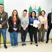 Seis pessoas estão alinhadas em pé, posando para a foto diante de um painel institucional do Governo do Estado do Rio Grande do Sul, que traz o brasão oficial no centro e as cores verde, vermelho e amarelo nas laterais. Da esquerda para a direita:

Um homem de óculos, cabelo curto escuro, veste blazer marrom, camisa preta e jeans.

Uma mulher de cabelos ondulados loiros, óculos e sorriso, veste blazer preto, blusa vinho e calça jeans larga.

Uma mulher de cabelos presos em coque alto, veste blusa cinza listrada e calça preta.

Uma mulher de cabelos escuros soltos, óculos, camisa azul e calça preta, segura papéis nas mãos.

Uma mulher de cabelos curtos castanhos, veste blazer rosa-claro, blusa estampada e calça marrom.

Um homem de barba curta, óculos, blazer preto, camiseta branca e calça escura.

Todos estão sorrindo e usam crachás de identificação, sugerindo um ambiente institucional ou oficial.