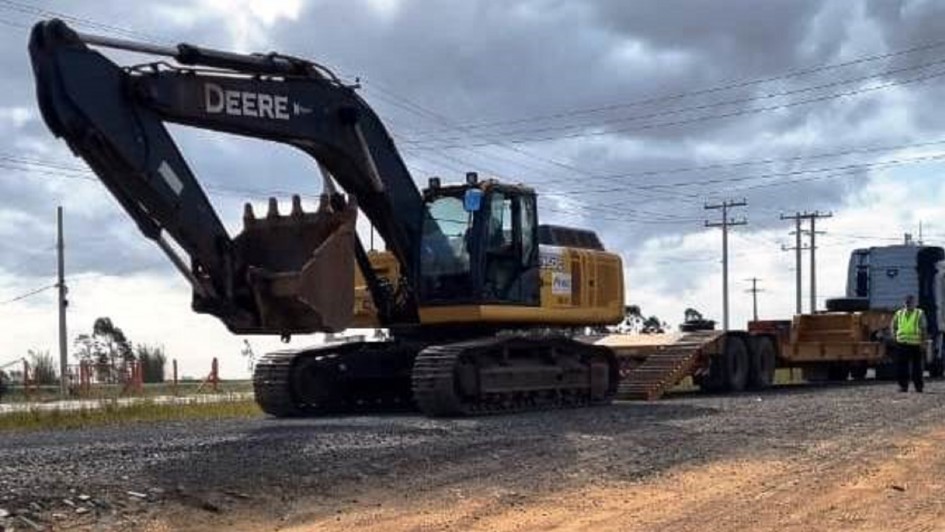 A foto mostra uma escavadeira de grande porte sendo transportada por caminhão prancha em trecho de obras viárias. O maquinário está sobre terreno de brita e areia, próximo à rodovia, com postes de energia ao fundo e céu nublado. Um trabalhador com colete refletivo acompanha a operação.