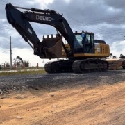 A foto mostra uma escavadeira de grande porte sendo transportada por caminhão prancha em trecho de obras viárias. O maquinário está sobre terreno de brita e areia, próximo à rodovia, com postes de energia ao fundo e céu nublado. Um trabalhador com colete refletivo acompanha a operação.