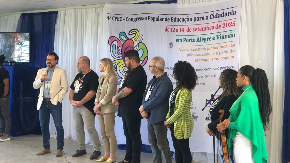 Fotografia tirada em ambiente interno durante o 4º Congresso Popular de Educação para a Cidadania, realizado entre os dias 12 e 14 de setembro de 2025. Nove pessoas estão em pé sobre um palco improvisado. À esquerda, um homem de blazer claro fala ao microfone. Ao fundo, há uma faixa com o nome e informações do evento, destacando os locais (Porto Alegre e Viamão) e o lema: "Vamos construir juntos políticas públicas criadas a partir do protagonismo popular". A decoração inclui tecidos brancos e azuis nas paredes e uma TV ao lado esquerdo, exibindo uma imagem de um homem com capacete. Entre os participantes no palco, há diversidade de gênero, etnia e trajes, reforçando o caráter inclusivo e comunitário do evento.