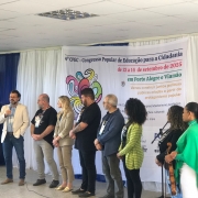 Fotografia tirada em ambiente interno durante o 4º Congresso Popular de Educação para a Cidadania, realizado entre os dias 12 e 14 de setembro de 2025. Nove pessoas estão em pé sobre um palco improvisado. À esquerda, um homem de blazer claro fala ao microfone. Ao fundo, há uma faixa com o nome e informações do evento, destacando os locais (Porto Alegre e Viamão) e o lema: "Vamos construir juntos políticas públicas criadas a partir do protagonismo popular". A decoração inclui tecidos brancos e azuis nas paredes e uma TV ao lado esquerdo, exibindo uma imagem de um homem com capacete. Entre os participantes no palco, há diversidade de gênero, etnia e trajes, reforçando o caráter inclusivo e comunitário do evento.