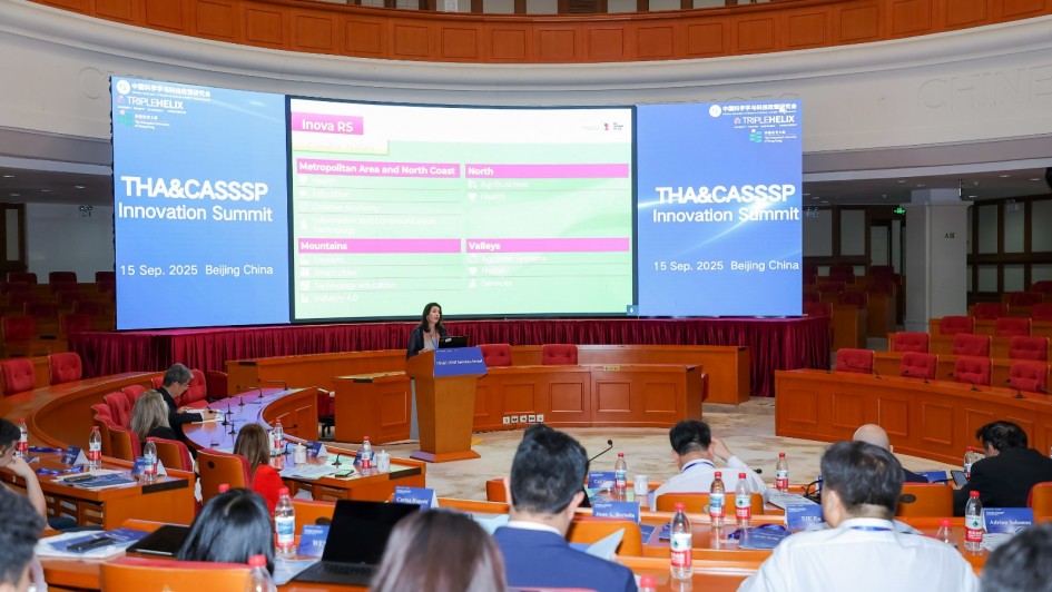 Auditório amplo com cadeiras dispostas em formato semicircular, onde ocorre o THA&CASSPS Innovation Summit, realizado em 15 de setembro de 2025, em Pequim, na China. No centro, uma palestrante apresenta no púlpito, enquanto dois grandes telões exibem informações sobre o evento e o programa Inova RS, com destaque para áreas como saúde, educação, economia criativa, tecnologia e turismo. Participantes acompanham a apresentação sentados, alguns com laptops e materiais de apoio sobre as mesas.