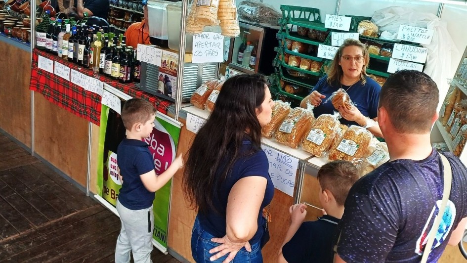 A imagem mostra uma cena movimentada de feira em um estande de venda de pães e produtos coloniais. No centro da imagem, uma mulher loira, vestida com camiseta azul, está atendendo uma família composta por dois adultos e duas crianças. Ela segura um pacote de pão e parece estar explicando algo sobre o produto. Sobre o balcão estão expostos diversos pacotes de pães, cucas e produtos similares, embalados em plástico transparente.

Ao fundo, prateleiras verdes estão cheias de mais pães e bolachas. Placas escritas à mão informam os preços dos produtos, como “PÃES $10,00”, “CUCA”, “BOLACHA $10,00”, “CANUDINHO $15,00”, e “PALITOS WAFFLE $8,00”. Outra placa no balcão diz “POR FAVOR NÃO APERTAR OS PÃES E CUCAS”.

À esquerda, outro estande vende vinhos, sucos e produtos artesanais, com garrafas expostas e utensílios de cerâmica pendurados. Há uma faixa vermelha xadrez decorando a banca. Crianças observam curiosas os produtos enquanto os pais conversam com a vendedora.

O ambiente é rústico, com piso de madeira, típico de feiras coloniais ou de produtos artesanais. A atmosfera transmite um clima familiar e acolhedor.