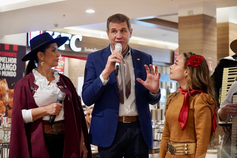 Close de três pessoas no palco do shopping. À esquerda, a cantora Anelise Severo, mulher com chapéu e capa grená segura microfone; ao centro, Ernani Polo, de terno azul e gravata estilizada, fala ao microfone; à direita, a cantora Luiza Barbosa, uma jovem com blusa marrom e lenço vermelho ouve atentamente. Ao fundo, a placa de uma lanchonete é visível.