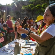 A imagem mostra um evento ao ar livre com crianças escoteiras, vestindo uniformes azuis com lenços verdes, observando uma mulher que segura e explica algo. Há mesas com livros e materiais educativos, além de várias pessoas ao fundo.