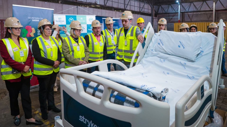Um grupo de pessoas com coletes refletivos amarelos e capacetes de segurança está reunido em um galpão. À frente, uma cama hospitalar nova, coberta com lençóis brancos estampados com o nome "Santa Casa". Os participantes observam atentamente a cama, posicionados ao redor dela. Faixas e banners institucionais da Secretaria da Saúde e da Santa Casa estão dispostos ao fundo. Entre o grupo, está a secretária da Saúde, Arita Bergmann.