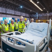 Um grupo de pessoas com coletes refletivos amarelos e capacetes de segurança está reunido em um galpão. À frente, uma cama hospitalar nova, coberta com lençóis brancos estampados com o nome "Santa Casa". Os participantes observam atentamente a cama, posicionados ao redor dela. Faixas e banners institucionais da Secretaria da Saúde e da Santa Casa estão dispostos ao fundo. Entre o grupo, está a secretária da Saúde, Arita Bergmann.