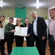 Foto em uma sala pintada em tons de verde. Quatro mulheres e dois homens posam para a foto. As duas mulheres ao centro seguram um documento assinado. Ao fundo, aparecem dois estudantes com uniformes na cor azul marinho. 
