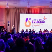 Fotografia de um auditório cheio durante a 6ª Conferência Estadual de Políticas para as Mulheres – Miguelina Vecchio. No palco, há uma mesa composta por diversas mulheres, atrás da qual está um grande telão com a identidade visual do evento. À esquerda, uma mulher fala no púlpito, enquanto fotógrafos registram o momento. À direita, há uma intérprete de Libras. A plateia, formada majoritariamente por mulheres, ocupa todas as cadeiras disponíveis. O ambiente está iluminado com luzes em tons de roxo e vermelho.