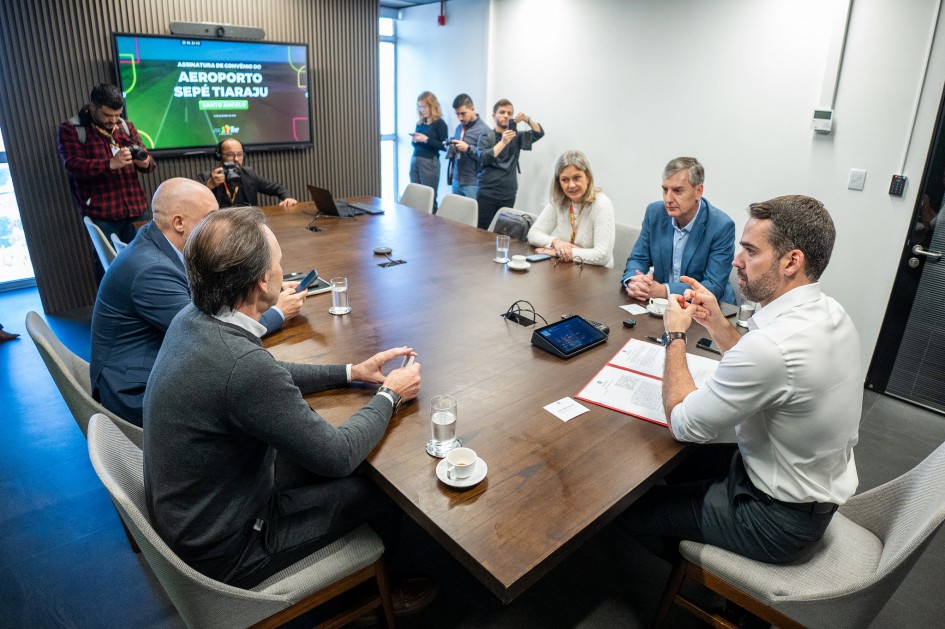 Em uma sala de reuniões, ao redor de uma mesa retangular, estão reunidos quatro homens e uma mulher. O governador está na ponta da mesa, gesticulando. Ao fundo, há um telão com uma apresentação sobre o Aeroporto Sepé Tiaraju.