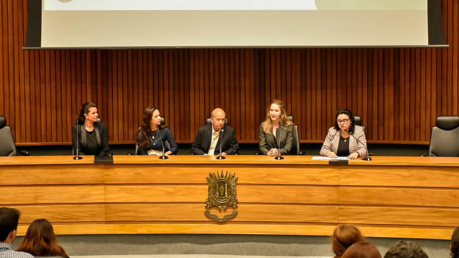 A imagem mostra uma mesa de conferência em um auditório com cinco pessoas sentadas atrás de uma bancada de madeira. Atrás delas, há um painel de madeira com ripas verticais e, acima, uma tela de projeção parcialmente visível com elementos gráficos. Da esquerda para a direita, vemos:
Uma mulher de cabelos escuros presos, vestida com roupas formais escuras.
Uma mulher de cabelos longos castanhos soltos, usando uma blusa azul-marinho e colar.
Um homem calvo, vestindo paletó escuro e camisa clara.
Uma mulher loira de cabelos soltos, usando blazer xadrez escuro.
Uma mulher de óculos, com cabelo escuro preso, vestindo blazer xadrez claro, que está lendo ou falando em um microfone, com papéis à sua frente.
Na frente da bancada de madeira há um brasão metálico fixado. Vemos também o público assistindo ao evento, com algumas cabeças visíveis na parte inferior da imagem. O ambiente sugere uma audiência ou seminário oficial, possivelmente em um contexto governamental ou institucional.