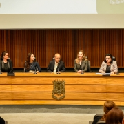 A imagem mostra uma mesa de conferência em um auditório com cinco pessoas sentadas atrás de uma bancada de madeira. Atrás delas, há um painel de madeira com ripas verticais e, acima, uma tela de projeção parcialmente visível com elementos gráficos. Da esquerda para a direita, vemos:
Uma mulher de cabelos escuros presos, vestida com roupas formais escuras.
Uma mulher de cabelos longos castanhos soltos, usando uma blusa azul-marinho e colar.
Um homem calvo, vestindo paletó escuro e camisa clara.
Uma mulher loira de cabelos soltos, usando blazer xadrez escuro.
Uma mulher de óculos, com cabelo escuro preso, vestindo blazer xadrez claro, que está lendo ou falando em um microfone, com papéis à sua frente.
Na frente da bancada de madeira há um brasão metálico fixado. Vemos também o público assistindo ao evento, com algumas cabeças visíveis na parte inferior da imagem. O ambiente sugere uma audiência ou seminário oficial, possivelmente em um contexto governamental ou institucional.
