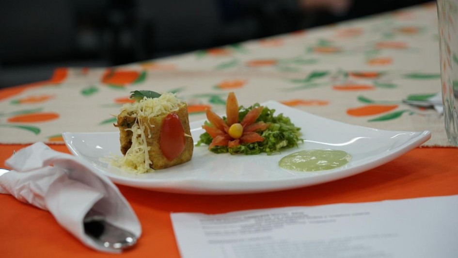 Um prato branco quadrado apresenta uma pequena porção de comida elaborada de forma decorativa. À esquerda, há um pedaço de torta salgada ou empada, coberta com queijo ralado e decorada com meio tomate cereja e uma folha verde. Ao centro, há uma guarnição de alface picada, enfeitada com fatias de tomate dispostas em forma de flor, com uma bolinha amarela no centro. À direita do prato, há uma pequena porção de molho claro esverdeado. O prato está sobre uma toalha de mesa com estampas de frutas laranja e verdes, e ao fundo há objetos desfocados.