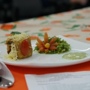 Um prato branco quadrado apresenta uma pequena porção de comida elaborada de forma decorativa. À esquerda, há um pedaço de torta salgada ou empada, coberta com queijo ralado e decorada com meio tomate cereja e uma folha verde. Ao centro, há uma guarnição de alface picada, enfeitada com fatias de tomate dispostas em forma de flor, com uma bolinha amarela no centro. À direita do prato, há uma pequena porção de molho claro esverdeado. O prato está sobre uma toalha de mesa com estampas de frutas laranja e verdes, e ao fundo há objetos desfocados.