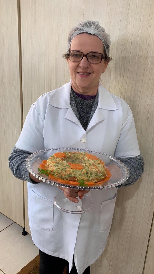Uma mulher sorri para a câmera segurando um prato de vidro redondo com bordas trabalhadas. Ela usa jaleco branco, malha cinza por baixo, touca de proteção nos cabelos e óculos de grau. No prato, há uma preparação culinária em formato de anel, feita com legumes picados, decorada com rodelas de cenoura e folhas de salsinha ao redor. O fundo mostra um armário de madeira clara.