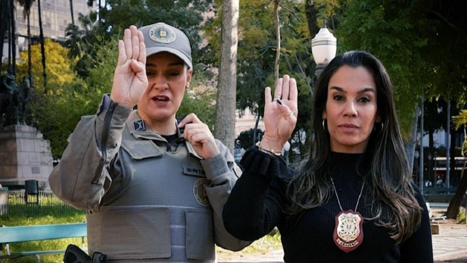 Duas mulheres fazem o sinal para identificar mulheres em situação de violência. Elas estão com a mão direita erguida, com o polegar dobrado sobre a mão e os quatro dedos para cima. 