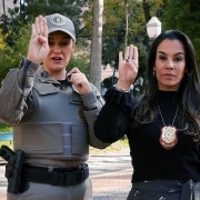 Duas mulheres fazem o sinal para identificar mulheres em situação de violência. Elas estão com a mão direita erguida, com o polegar dobrado sobre a mão e os quatro dedos para cima. 