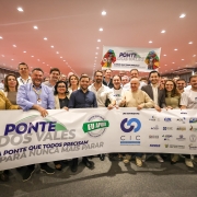 Um grupo numeroso de pessoas posa sorridente em um auditório, segurando uma grande faixa branca com a inscrição em destaque: “Ponte dos Vales – A ponte que todos precisam para nunca mais parar”. No centro da faixa aparecem os logotipos do movimento liderado pela CIC Vale do Taquari e de diversas entidades apoiadoras, como FIERGS, Sistema Farsul, Fecomércio RS e Federasul, entre outras. Algumas pessoas vestem camisetas brancas com o mesmo slogan da campanha. Ao fundo, outra faixa pendurada reforça a identidade do movimento, decorada com a imagem de uma ponte e uma bandeira do Rio Grande do Sul estilizada. O ambiente é iluminado com luzes de teto, e todos aparentam estar em clima de união e apoio ao projeto.