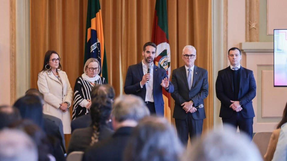 Cinco pessoas em pé diante de uma platéia; a do centro fala ao microfone. Atrás, bandeiras do Brasil e outra com vermelho, verde e branco. Ambiente formal, tipo conferência.

