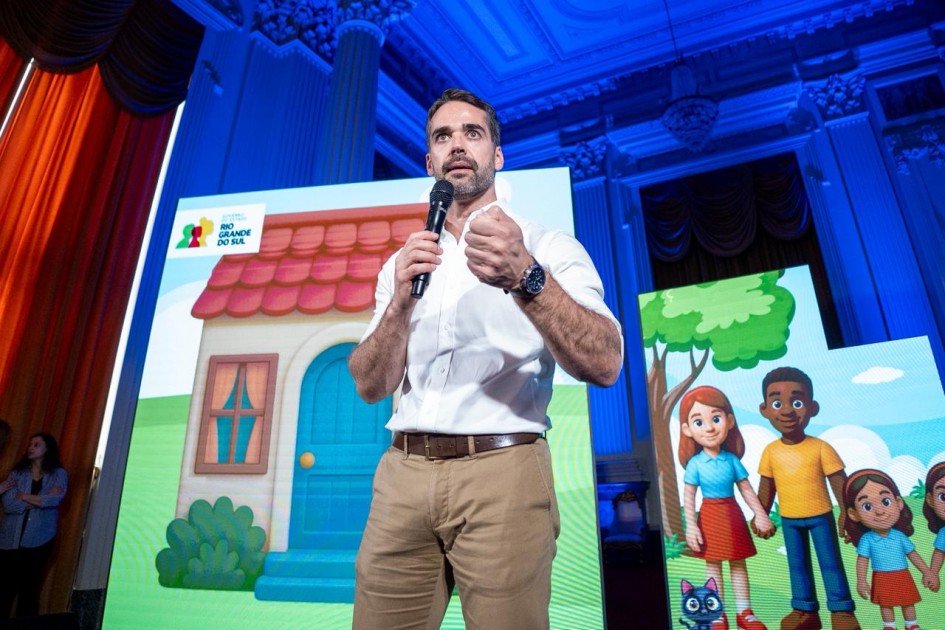 O governador Eduardo Leite aparece em primeiro plano, usando camisa branca de mangas dobradas e calça bege. Ele segura um microfone e faz um gesto com a mão, enquanto fala. Ao fundo, um telão exibe uma ilustração colorida com uma casa e personagens infantis.