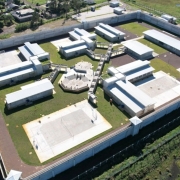 Visão aérea de uma instalação, possivelmente uma prisão ou complexo de segurança. O layout apresenta várias construções interligadas por passarelas, com cercas altas ao redor. Também podemos ver uma área de esporte ao fundo, provavelmente destinada à recreação dos internos. O design parece organizado e separado em várias seções, com estruturas de controle de segurança visíveis, como torres e muros. Há um espaço aberto com grama e alguns prédios adicionais ao fundo, sugerindo uma área ampla.