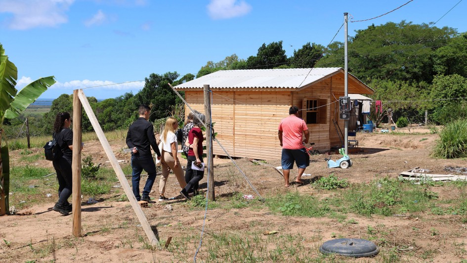 Seis pessoas caminham em direção a uma pequena casa de madeira recém-construída, situada em uma área rural. A casa tem telhado de telhas brancas, janelas abertas e está cercada por vegetação baixa e algumas árvores ao fundo. O grupo é formado por homens e mulheres, alguns carregando mochilas e documentos. O terreno é de terra batida com pontos de grama e objetos espalhados, como um pneu e ferramentas. À direita da casa, há roupas estendidas em um varal e, próximo à parede, um medidor de energia elétrica fixado em um poste. O céu está azul com algumas nuvens.