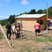 Seis pessoas caminham em direção a uma pequena casa de madeira recém-construída, situada em uma área rural. A casa tem telhado de telhas brancas, janelas abertas e está cercada por vegetação baixa e algumas árvores ao fundo. O grupo é formado por homens e mulheres, alguns carregando mochilas e documentos. O terreno é de terra batida com pontos de grama e objetos espalhados, como um pneu e ferramentas. À direita da casa, há roupas estendidas em um varal e, próximo à parede, um medidor de energia elétrica fixado em um poste. O céu está azul com algumas nuvens.