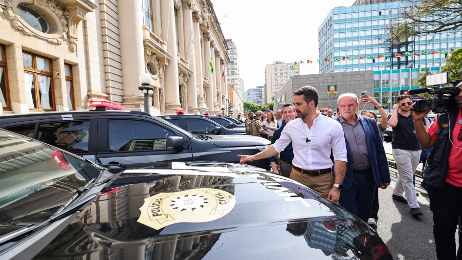 O governador Eduardo Leite aparece em frente ao Palácio Piratini, acompanhado de outras autoridades e assessores. Ele veste camisa branca e calça bege, caminhando ao lado de viaturas da Polícia Civil estacionadas na rua. Há também imprensa registrando o momento.
