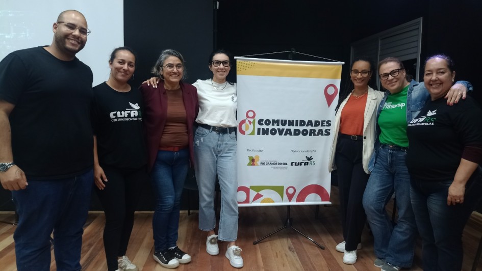 A imagem mostra um grupo de sete pessoas posando para uma foto em frente a um banner com o nome "COMUNIDADES INOVADORAS". O banner também inclui logos de organizações apoiadoras. As pessoas estão sorrindo e se vestem de forma casual, com camisetas e roupas variadas. O evento parece estar relacionado ao empreendedorismo ou qualificação, dado o contexto do banner e o ambiente de encontro. O fundo é de um espaço fechado, com piso de madeira e iluminação suave.