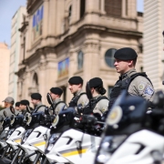 Policiais militares da Brigada Militar estão perfilados em frente a uma fileira de motos oficiais, durante cerimônia em Porto Alegre. Os agentes vestem uniforme cinza, coletes pretos e boinas pretas, permanecendo em posição de atenção. As motocicletas brancas estão alinhadas lado a lado, com faixas amarelas e pretas. Ao fundo, aparecem prédios históricos do centro da cidade, incluindo uma igreja com vitrais coloridos visíveis em sua fachada.