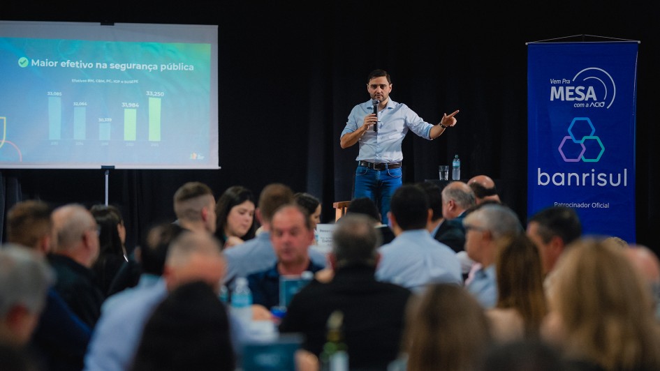 Gabriel aparece de camisa azul clara e calça jeans e está em pé no palco, falando ao microfone diante de várias pessoas sentadas. Ao fundo, há uma tela com gráfico sobre segurança pública e um banner azul do Banrisul.